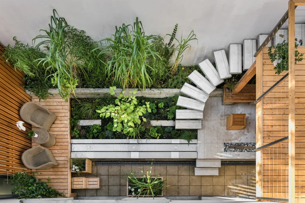 Aerial view of a modern urban garden with layered landscaping, featuring a wooden deck with two wicker chairs, lush greenery, and a spiral staircase, highlighting efficient use of small outdoor spaces.