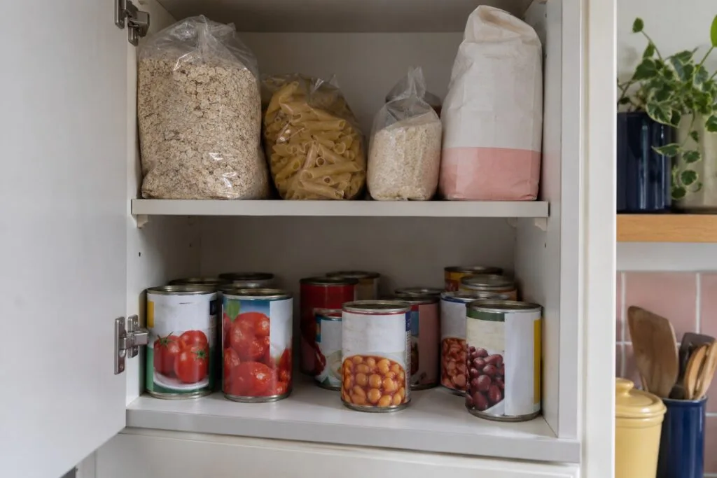 An open kitchen cupboard containing oats, pasta, rice, flour and tins of tomatoes and beans.