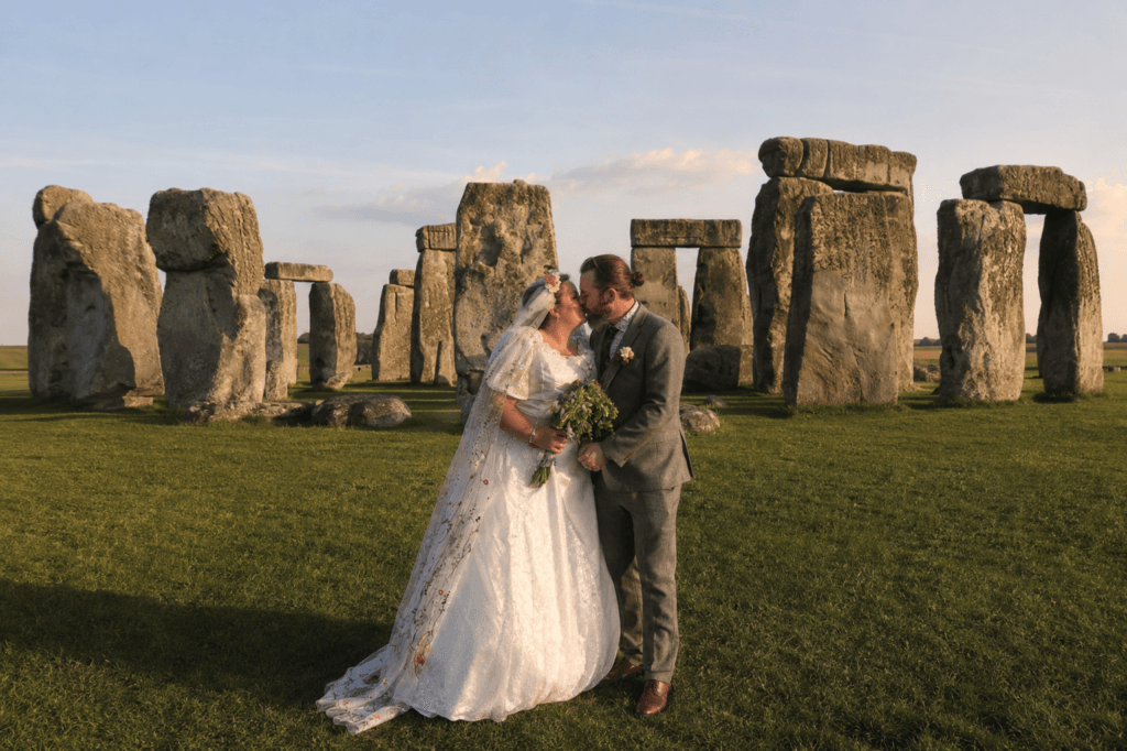 Niki and G sharing a kiss in their wedding clothes beside ancient standing stones